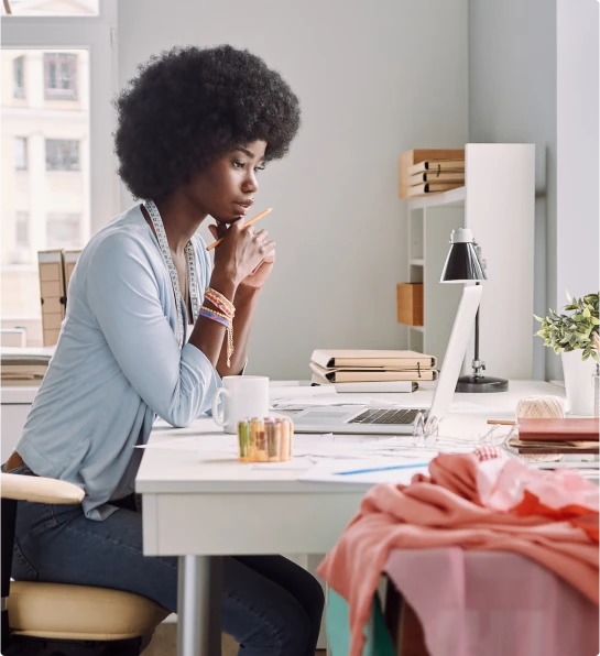 Mulher pensativa com cabelo afro, usando roupas casuais e trabalhando em seu laptop em um escritório com materiais de costura.