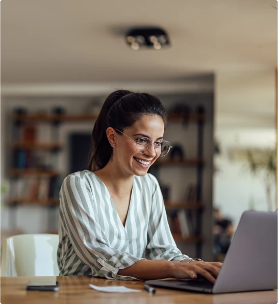 Mulher sorridente com óculos e blusa listrada, trabalhando em um notebook em um ambiente moderno e iluminado.