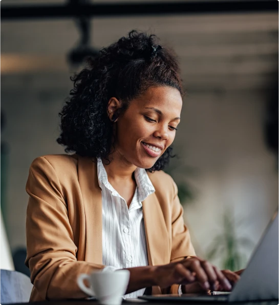Mulher sorridente com blazer bege, digitando em um laptop em um ambiente profissional com uma xícara de café.